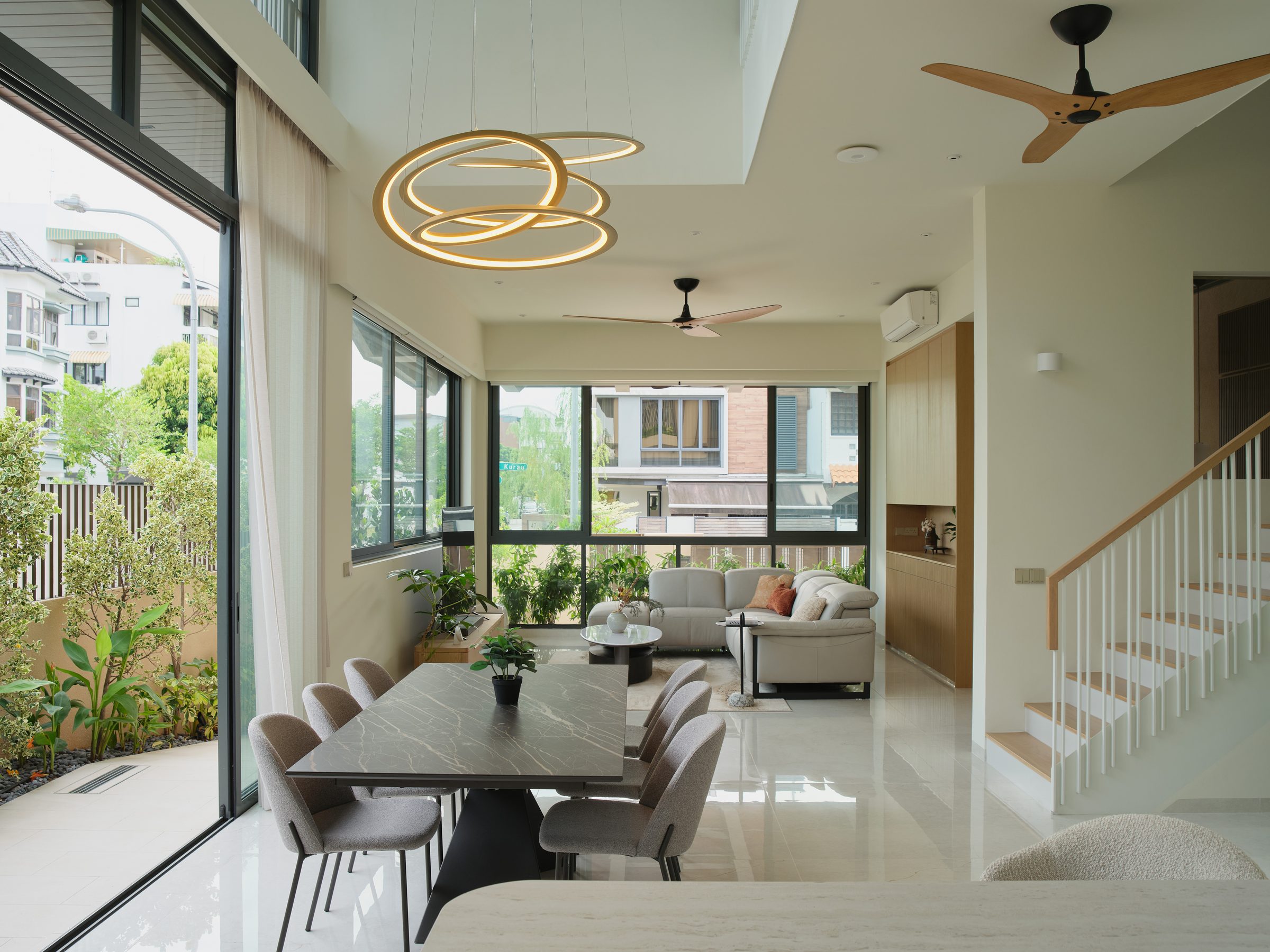 Double-volume dining space with a stone-topped table under a sculptural ring pendant, opening to the garden on one side and the living area beyond