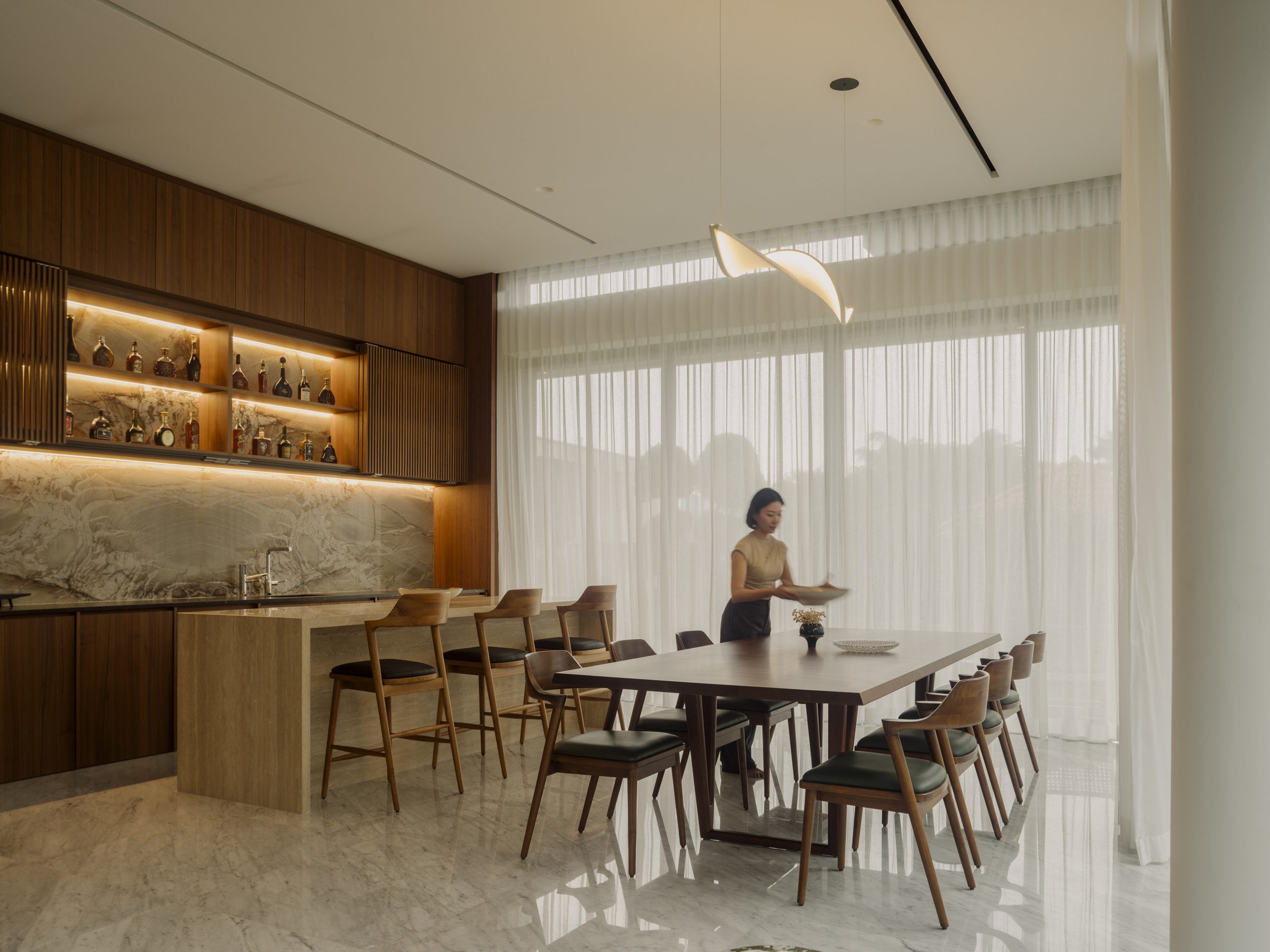 Dining table and island kitchen under a sculptural pendant, with sheer curtains drawn along the garden glazing