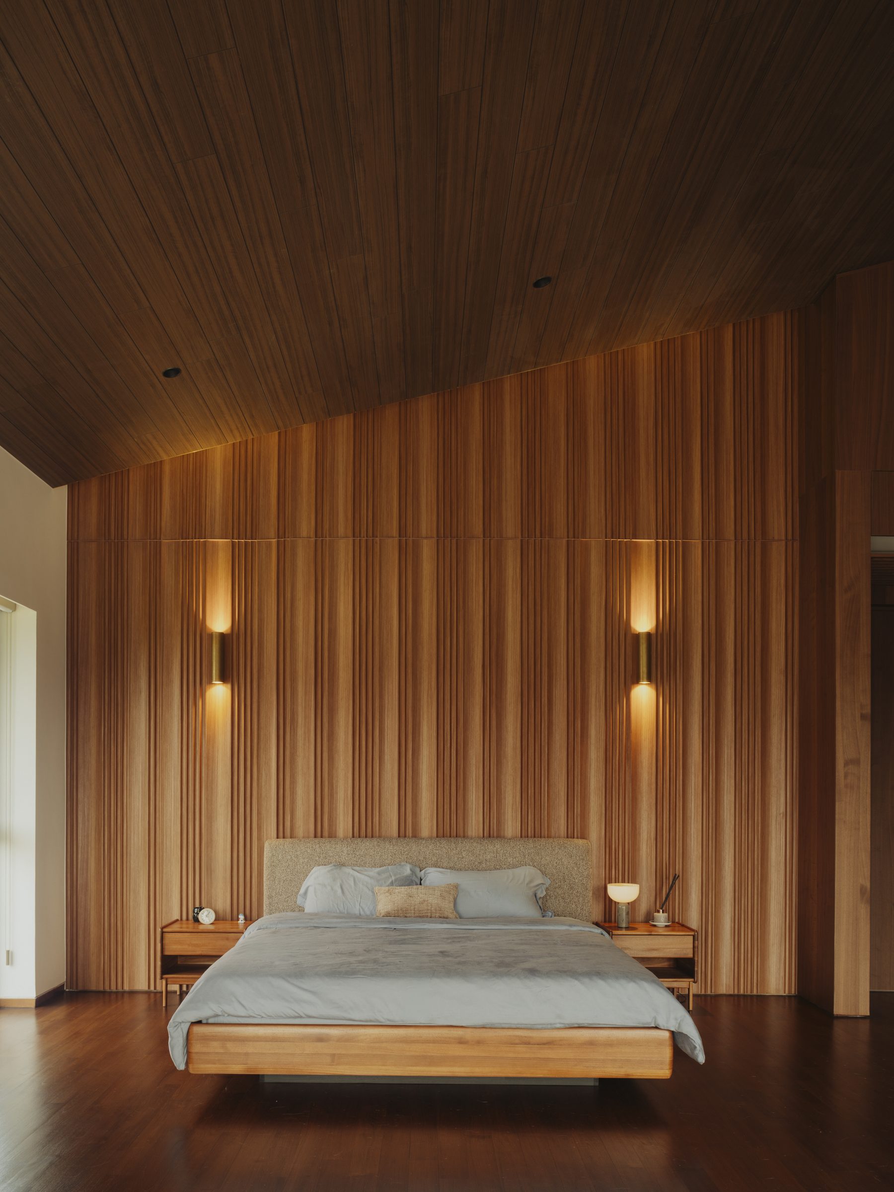 Master bedroom lined in timber, the bed set between wall lights under a raked ceiling